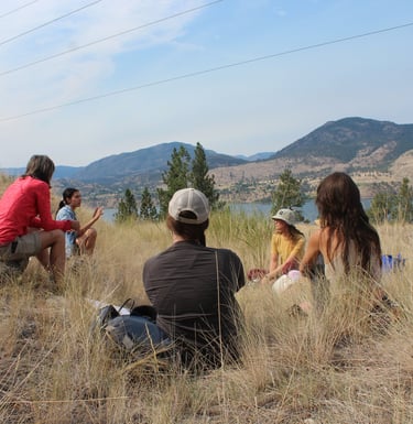 A group of people sitting on a hill top with Skaha Lake and rolling mountains in the background.