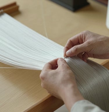 A photograph focusing on the hands of a Japanese artisan carefully weaving silk threads in a minimalist, sun-drenched Kyoto studio. The scene is calm and elegant. Colors include soft silver sage and warm cream.