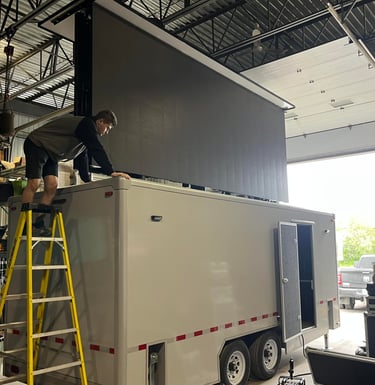 A technician installs a large mobile LED screen onto a silver event trailer inside a warehouse.