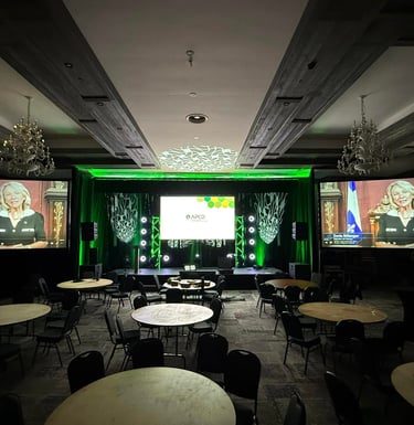 Professional conference room set with stage lighting, round banquet tables, and large screens for a live presentation.