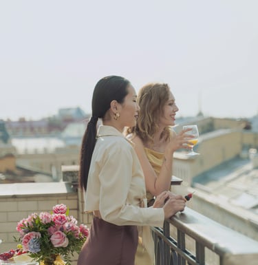 Woman with pink hair working remotely on a laptop in a modern hotel room with mountain views, next to a purple suitcase.