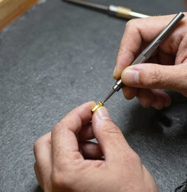 A master jeweler's hands working with precision tools on a gold setting. The scene is shot from above on a dark slate grey workbench with warm cream light focusing on the meticulous work.