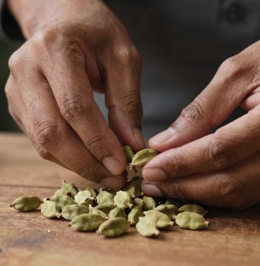 Close-up of a South Asian / Indian spice expert's hands carefully grading fresh green cardamom pods on a rustic wooden table. The lighting is soft and natural, emphasizing trust and quality control.