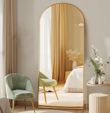 Gold arched floor mirror leaning against a wall in a modern bedroom with a green velvet chair.