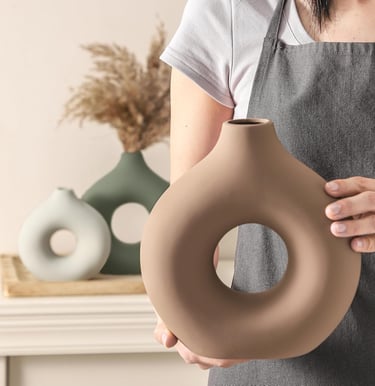 A person in a gray apron holding a modern brown ceramic donut vase with a matte finish.