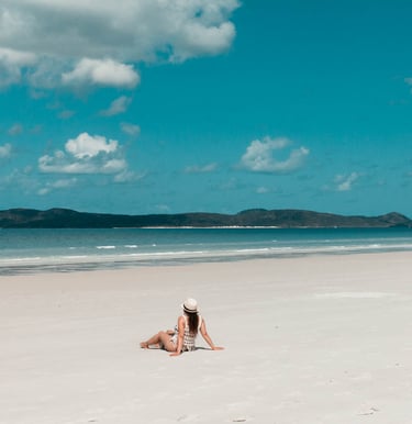 A woman wearing a sun hat sits on a white sand tropical beach with blue ocean water and a cloudy sky.