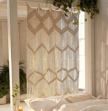 A decorative boho macrame wall hanging used as a stylish room divider in a bright, sunlit bedroom.
