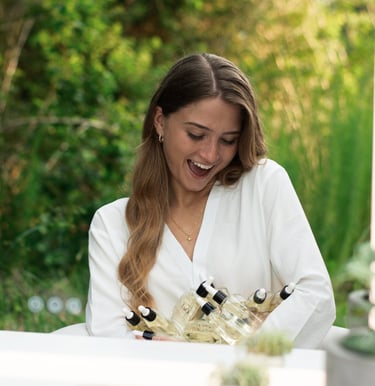 Smiling woman in a white robe holding several organic facial oil serum bottles in a lush garden setting.