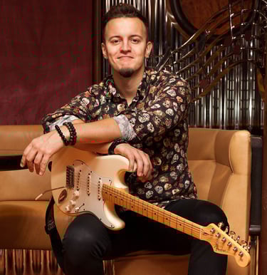 Guitarist Lucas Vianna sitting on a couch with his white Fender Stratocaster guitar on his lap, wearing a colorful shirt and 