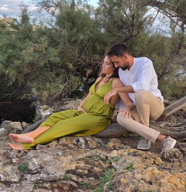 A family of three sitting on rocky terrain near a river with greenery in the background, enjoying an outdoor moment.