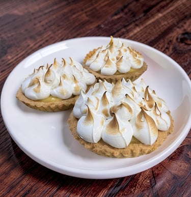 Three homemade mini lemon meringue tarts with toasted peaks on a white plate over dark wood.