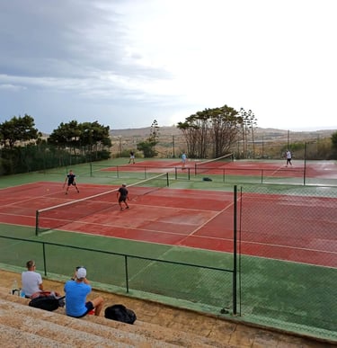 People playing tennis on outdoor red clay courts with scenic landscape views under a cloudy sky.