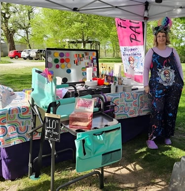 A table set up at a festival, colorful paints displayed with Sarah kipper in front of table smiling 