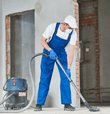 Trabajador con mono de tirantes azul, camiseta blanca y gorra, limpiando en una obra con aspirador.