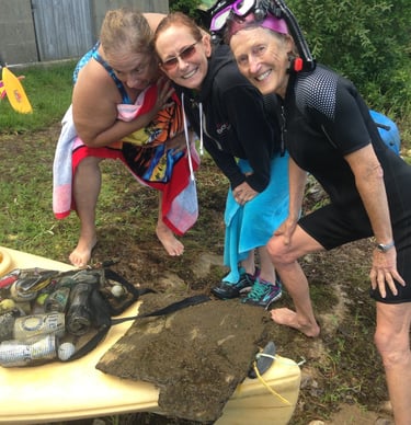 Susan Baur posed with two other women next to a kayak and a big of trash. All are in swimming gear. 