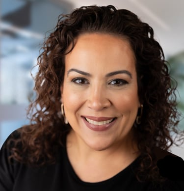 Professional headshot of a smiling woman with curly brown hair wearing a black shirt and hoop earrings.
