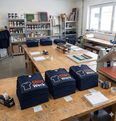 Stacks of printed navy blue t-shirts on a wooden table in a professional screen printing workshop.