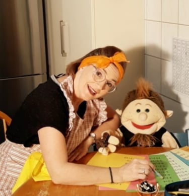 Betti Konfetti mit Handpuppe am Küchentisch erklärt die Schulaufgaben.