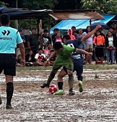 Soccer players competing for a ball on a muddy field during a local match with spectators.