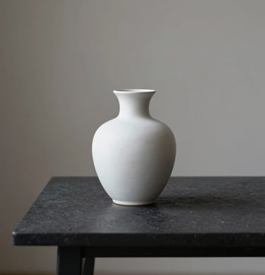 Minimalist still life of a single white ceramic vase on a dark charcoal table, soft side lighting, elegant Northern European home interior.