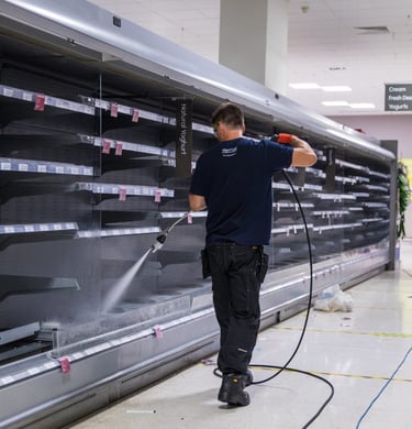 A maintenance worker pressure washing empty refrigerated shelves in a supermarket aisle.