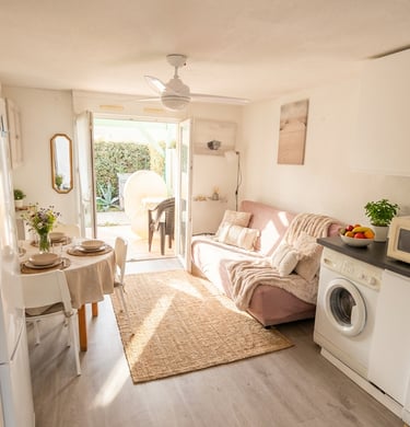 Bright studio apartment with a cozy pink sofa, dining table, and kitchen area leading to a sunny outdoor patio.