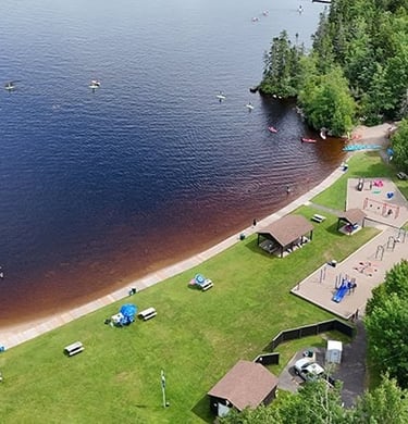 Aerial view of a public beach park with a grassy picnic area, playground, and people kayaking on the lake.