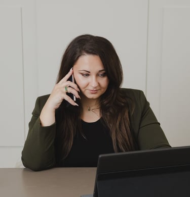Adriana in olive blazer talking on a smartphone while working on a tablet laptop.