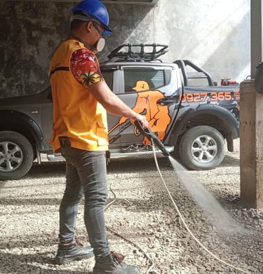 A worker using a high-pressure power washer to clean a gravel surface near a pickup truck.