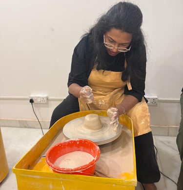 A woman wearing a yellow apron uses a pottery wheel to shape clay during a pottery workshop.