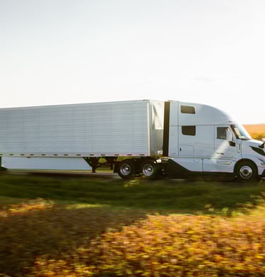 White semi-truck with a long trailer drives on a rural highway during golden hour, transporting goods.