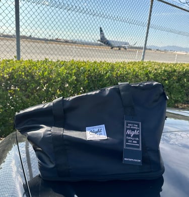 Black Night Supply Co. duffel bag with logo and hang tag, placed near airport fence