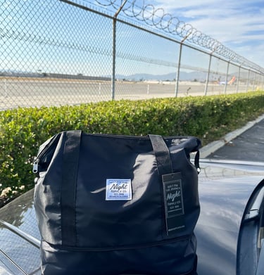 Black Night Supply Co. duffel bag with logo and hang tag, placed near airport fence