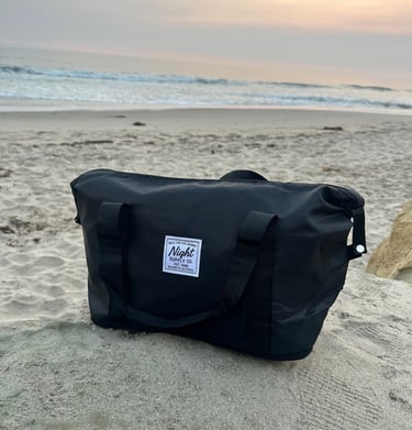 Black Night Supply Co. duffel bag on beach sand with sunset sky in background