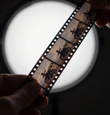 A close-up shot of a hand holding a strip of black and white film negatives up to a soft grey light source, revealing the frames of a motorcycle trip. Global / Western darkroom setting.