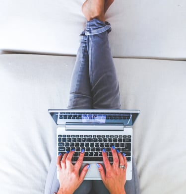 Femme en train de pianoter sur son ordinateur portable et qui a les jambes allongées sur le canapé blanc