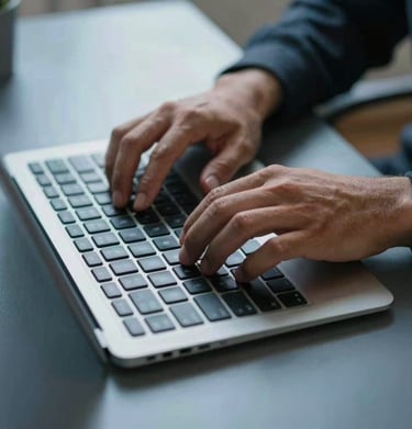 A close-up photograph of a North American / US person's hands typing on a sleek wireless keyboard on a clean steel blue desk.