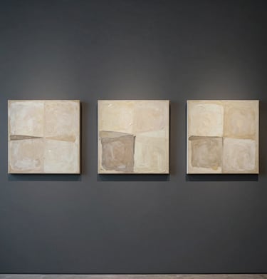 A triptych of three small, square abstract paintings in cream and taupe tones, arranged symmetrically on a dark grey charcoal wall in a contemporary space.