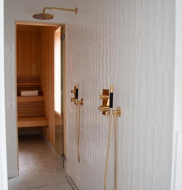 Modern luxury walk-in shower with gold fixtures and white subway tile next to a wood-paneled home sauna.