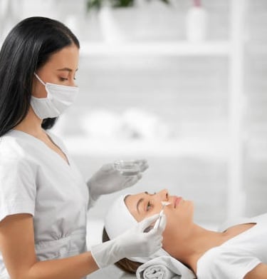 Professional esthetician in a mask applying a chemical peel facial treatment to a client in a spa.