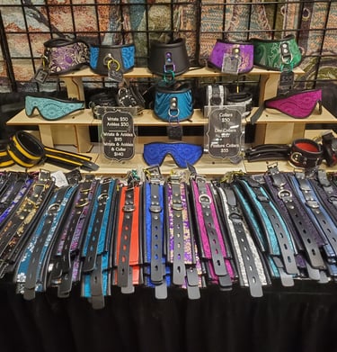 A display of colorful leather BDSM collars, wrist cuffs, and lace eye masks at a craft fair.