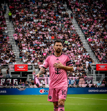 Lionel Messi in a pink Inter Miami jersey walking on the pitch in front of a crowded stadium.