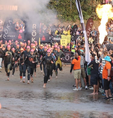 Triathletes in wetsuits start Ironman Wales swim on a beach with fire effects and a cheering crowd.