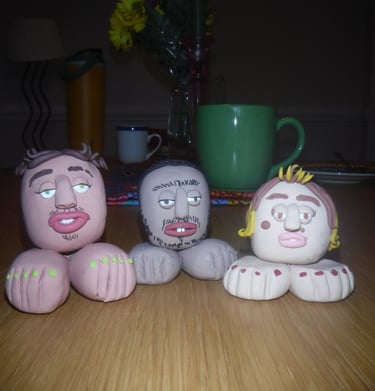 Three whimsical handmade clay figures with exaggerated facial features and feet on a wooden table.