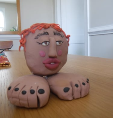 Handmade clay sculpture of a person's head with orange hair sitting on two large feet.