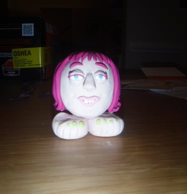 Handmade clay figurine of a pink-haired face resting on white feet with green toes.