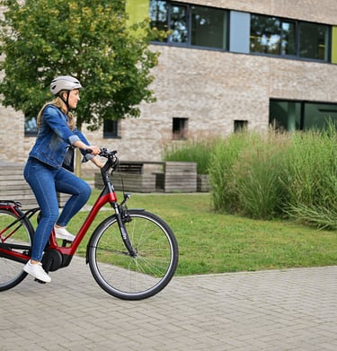 A woman wearing a helmet rides a red electric commuter bike on a paved path near a modern building.