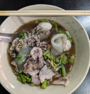A bowl of Thai boat noodles with sliced pork, fish balls, and morning glory in a rich savory broth.