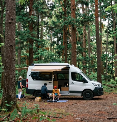 A rustic wooden mobile home is parked at the side of a quiet street with minimal traffic. The home has a triangular roof adorned with colorful pennant flags, and there are small windows on the back. The street is lined with traditional buildings and a stone wall, and the sky is partly cloudy, indicating a sunny day.
