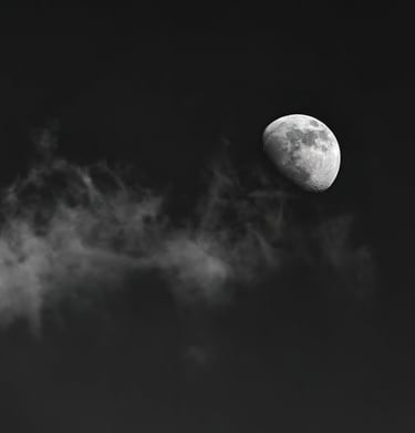 A black-and-white photograph of a line of cloud leading up to a gibbous moon.